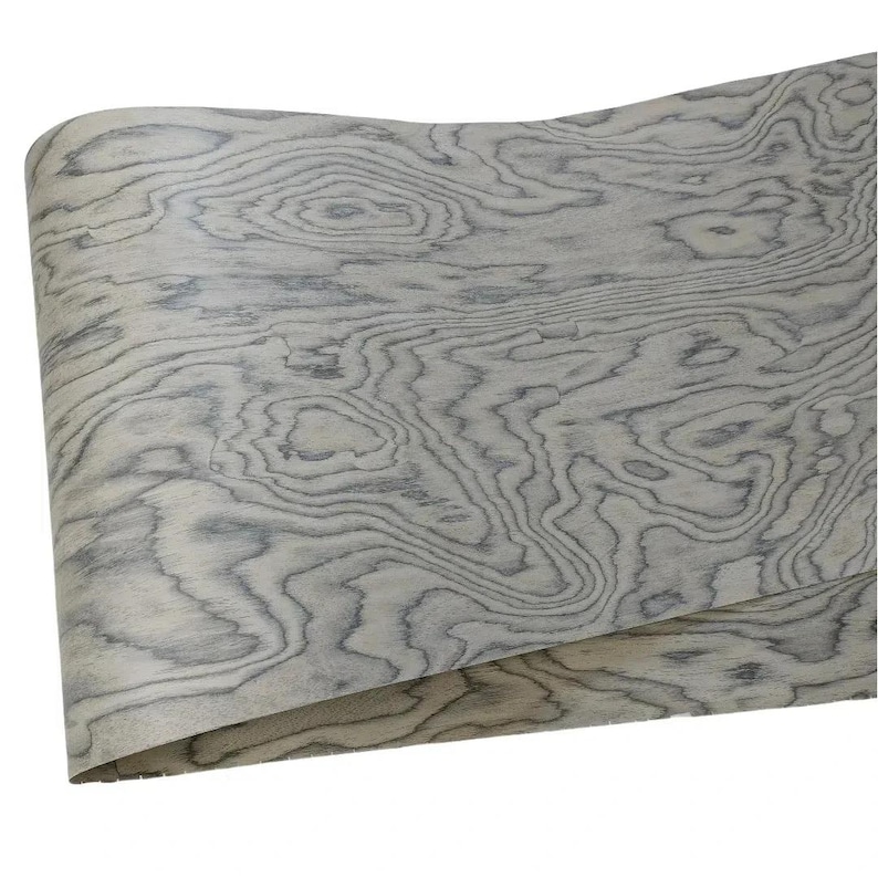 Puede incluir: Una l&aacute;mina enrollada de chapa de madera con un patr&oacute;n de vetas de madera gris claro y gris azulado. La chapa tiene un aspecto natural y org&aacute;nico con l&iacute;neas y nudos arremolinados.