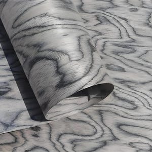 Puede incluir: Primer plano de un rollo de chapa de madera con un patr&oacute;n de vetas gris y blanco. La veta de la madera tiene un dise&ntilde;o ondulado y org&aacute;nico. La chapa est&aacute; parcialmente enrollada, revelando la parte inferior. El efecto general es una superficie natural y texturizada.