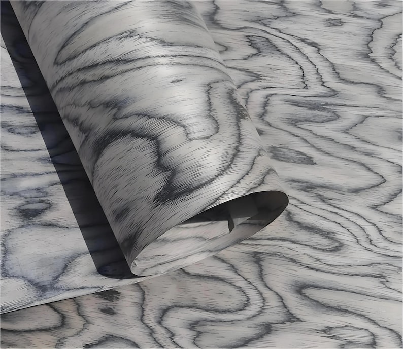 Puede incluir: Primer plano de un rollo de chapa de madera con un patr&oacute;n de vetas gris y blanco. La veta de la madera tiene un dise&ntilde;o ondulado y org&aacute;nico. La chapa est&aacute; parcialmente enrollada, revelando la parte inferior. El efecto general es una superficie natural y texturizada.