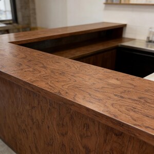 K&ouml;nnte beinhalten: Eine ma&szlig;gefertigte Holzbar mit einem satten, warmbraunen Finish. Die Bar verf&uuml;gt &uuml;ber eine Arbeitsplatte mit detailliertem Holzmaserungsmuster und einen erh&ouml;hten R&uuml;ckbereich. Das Design umfasst einen vertieften Bereich, der auf einen Arbeits- oder Lagerbereich hindeutet. Die Konstruktion der Bar ist robust und gut verarbeitet.