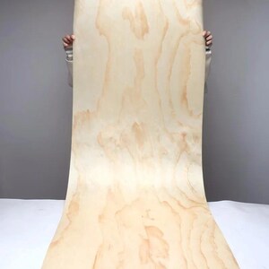 May include: A large sheet of light-colored plywood with a natural wood grain pattern. The wood has a warm, beige tone with darker brown streaks and swirls. The sheet is held up against a gray background.