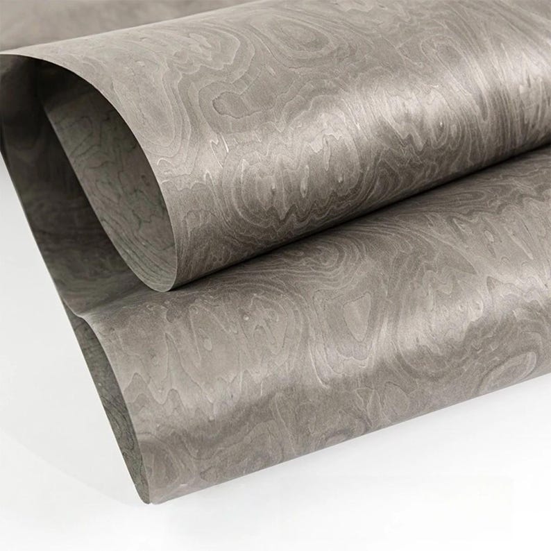 May include: A close-up of a rolled sheet of gray wood veneer. The veneer has a natural wood grain pattern with swirling lines and variations in tone. The edges of the sheet are visible, showing the thickness and flexibility of the material. The overall appearance is smooth and elegant.