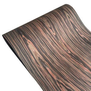 May include: A rolled sheet of wood veneer with a distinctive grain pattern. The veneer displays alternating bands of dark grey, reddish-brown, and light tan, creating a natural, textured appearance. The wood grain flows in a wavy, linear direction.