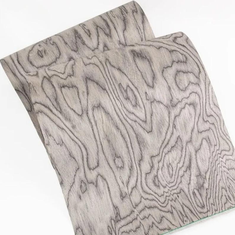 K&ouml;nnte beinhalten: Zwei Holzfurnierplatten mit einem grau-schwarzen Maserungsmuster. Die Holzmaserung hat ein wirbelndes, organisches Design. Die Platten sind leicht gebogen und zeigen die Textur und Flexibilit&auml;t des Materials. Das Holz hat ein nat&uuml;rliches, verwittertes Aussehen.