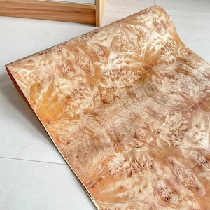 May include: A roll of wood veneer with a complex, swirling grain pattern in shades of light and dark brown. The veneer is partially unrolled, revealing its intricate design and texture. The wood grain has a natural, organic appearance.