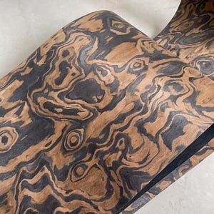 May include: Close-up of a roll of wood veneer with a striking pattern. The veneer features a natural wood grain in shades of brown and black, creating a marbled effect. The wood grain swirls and curves, adding visual interest to the surface.