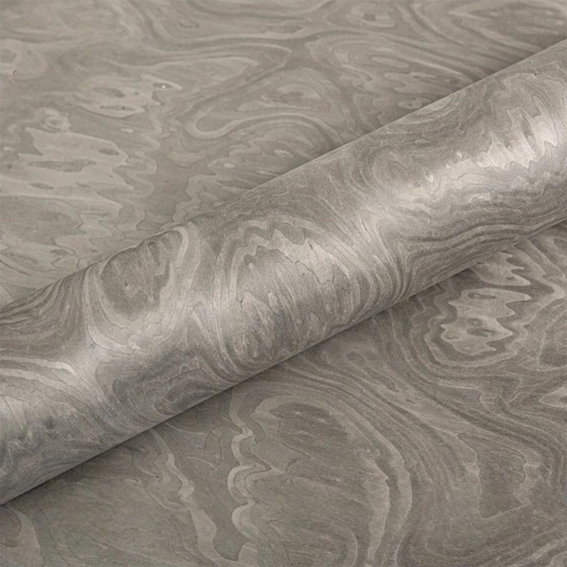 May include: A roll of gray marbled paper with a swirling pattern. The paper has a smooth, glossy finish and a subtle variation in tone, creating a sense of depth and texture. The paper is partially unrolled, revealing the pattern.