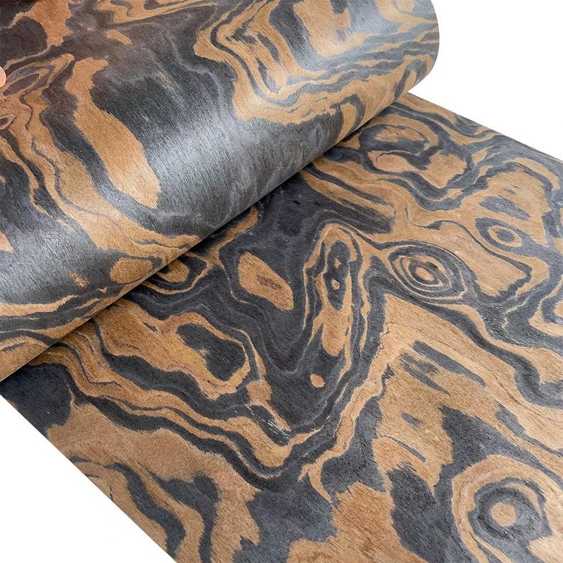 May include: Close-up of a roll of wood veneer with a swirling pattern. The veneer features a combination of dark gray and warm brown tones, creating a natural, organic aesthetic. The wood grain is visible, adding texture and depth to the material.