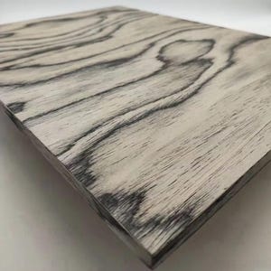 May include: A rectangular wooden panel with a light grey base and dark grey wood grain patterns. The wood grain has a natural, flowing design. The panel's edges are visible, showing the thickness of the wood.