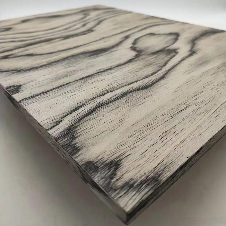 May include: A rectangular wooden panel with a light grey base and dark grey wood grain patterns. The wood grain has a natural, flowing design. The panel's edges are visible, showing the thickness of the wood.