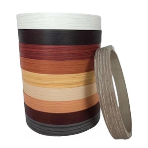 Puede incluir: Una pila de rollos de canto de borde en varios colores de vetas de madera, incluyendo blanco, negro y tonos de marrón y beige. Un solo rollo se encuentra de pie junto a la pila, mostrando la textura y el diseño del canto. Ideal para proyectos de carpintería y mobiliario.
