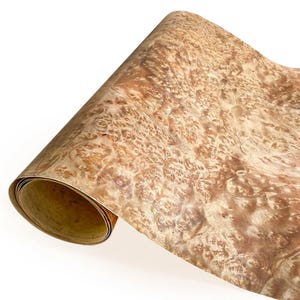 Könnte beinhalten: Eine gerollte Holzfurnierplatte mit einem komplexen, wirbelnden Maserungsmuster. Das Furnier zeigt eine warme, natürliche Farbpalette aus hellen und mittleren Brauntönen mit einem strukturierten, maserartigen Aussehen. Die Kanten der Rolle zeigen ein kontrastierendes, helleres Innere.