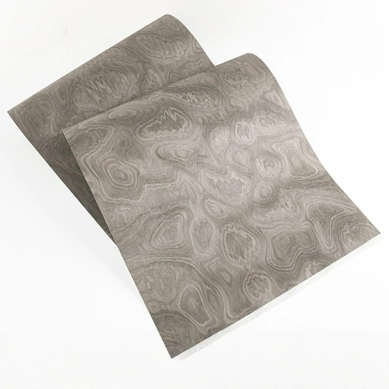 May include: Two sheets of gray decorative paper with a swirling wood grain pattern. The paper has a light gray base color with darker gray and white swirling patterns. The sheets are slightly overlapping, with the edges visible.