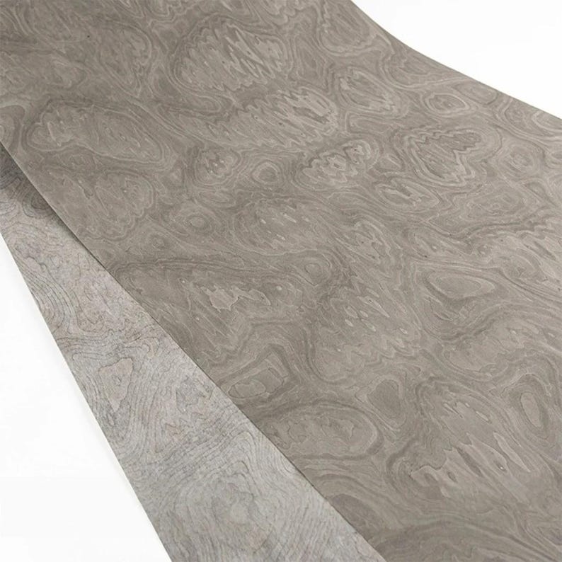 May include: A sheet of gray wood veneer with a swirling, organic grain pattern. The veneer has a light, neutral color and a smooth surface. The wood grain creates a natural, flowing design, suitable for furniture or decorative projects.