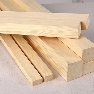 May include: Several unfinished, light-colored wooden beams and planks are arranged on a light-colored surface. The wood grain is visible, and the pieces vary in size and shape, suggesting they are for a construction or craft project.