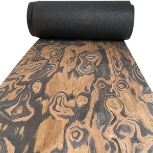May include: A roll of black material sits atop a sheet of wood with a unique grain pattern. The wood features swirling patterns in shades of brown and gray, creating a natural, organic aesthetic. The rolled material appears to be a dark, solid color.