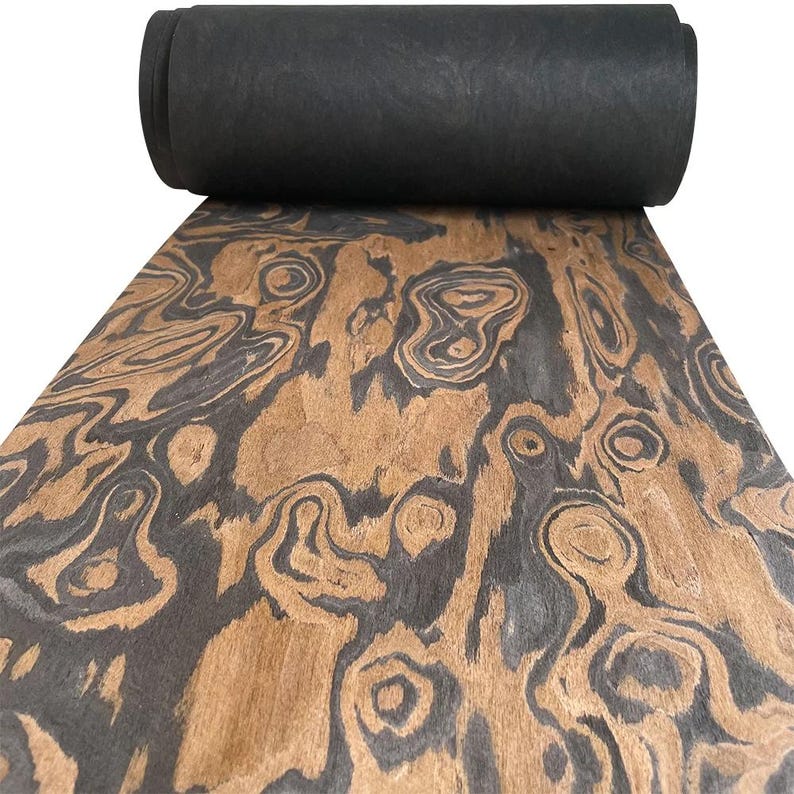 May include: A roll of black material sits atop a sheet of wood with a unique grain pattern. The wood features swirling patterns in shades of brown and gray, creating a natural, organic aesthetic. The rolled material appears to be a dark, solid color.