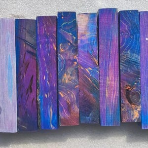 May include: Ten rectangular wooden blocks, each with a unique pattern of swirling blues, purples, and hints of orange. The wood grain is visible, adding texture to the vibrant, dyed surfaces. The blocks are arranged vertically, side by side.