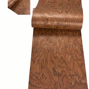 K&ouml;nnte beinhalten: Eine Rolle Holzfurnier mit einem satten, rotbraunen Farbton und einem auff&auml;lligen, wirbelnden Maserungsmuster. Das Furnier ist teilweise abgerollt und zeigt die komplizierten Details des nat&uuml;rlichen Designs des Holzes. Die Holzmaserung weist dunkle, wellenf&ouml;rmige Linien auf.