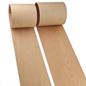 Può includere: Due rotoli di impiallacciatura in legno marrone chiaro, parzialmente srotolati, che mostrano la venatura del legno. L'impiallacciatura è di colore marrone chiaro e sembra sottile e flessibile. Lo sfondo è bianco.