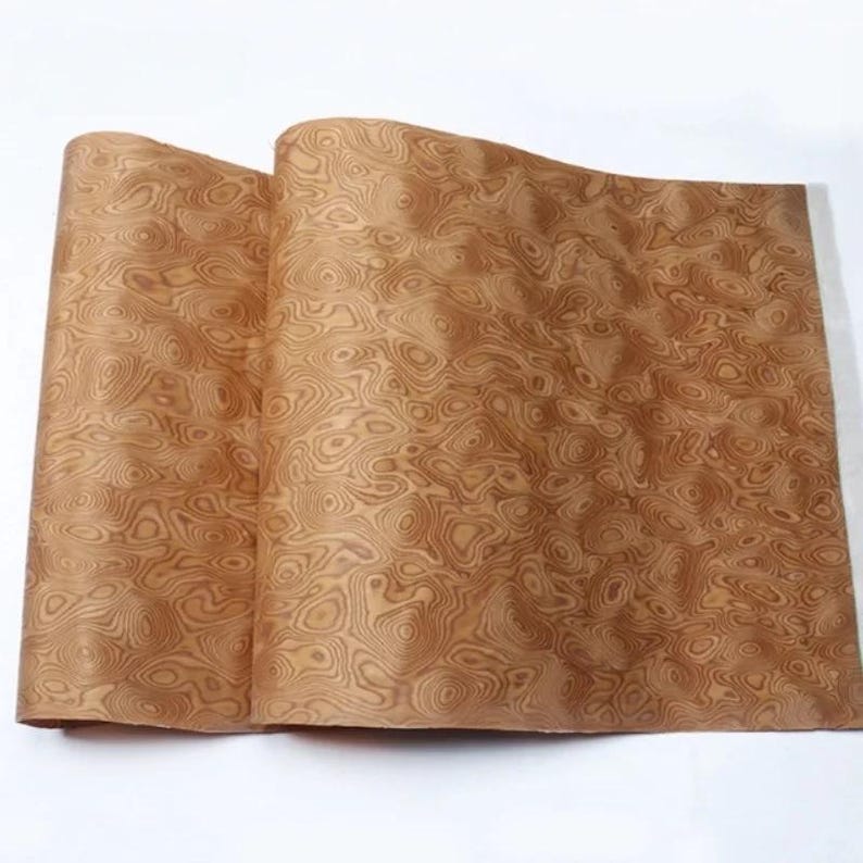 May include: Two sheets of wood veneer with a swirling, organic grain pattern in shades of brown and tan. The veneer is slightly curled, revealing the underside and texture of the material. The wood grain creates a natural, flowing design.