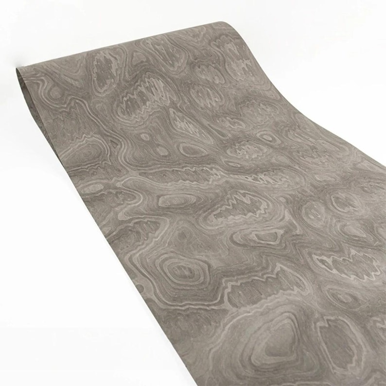May include: A rolled-up sheet of gray decorative paper with a wood grain pattern. The paper has a swirling, organic design in varying shades of gray and white, creating a textured appearance. The paper is against a white background.