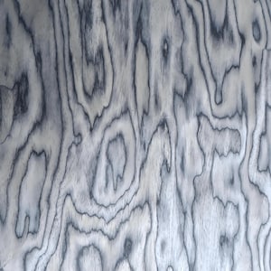 May include: Close-up of a wood grain pattern with a swirling, organic design. The wood features a light grey base with dark grey, almost black, swirling lines that create a textured appearance. The grain pattern is consistent throughout the image.