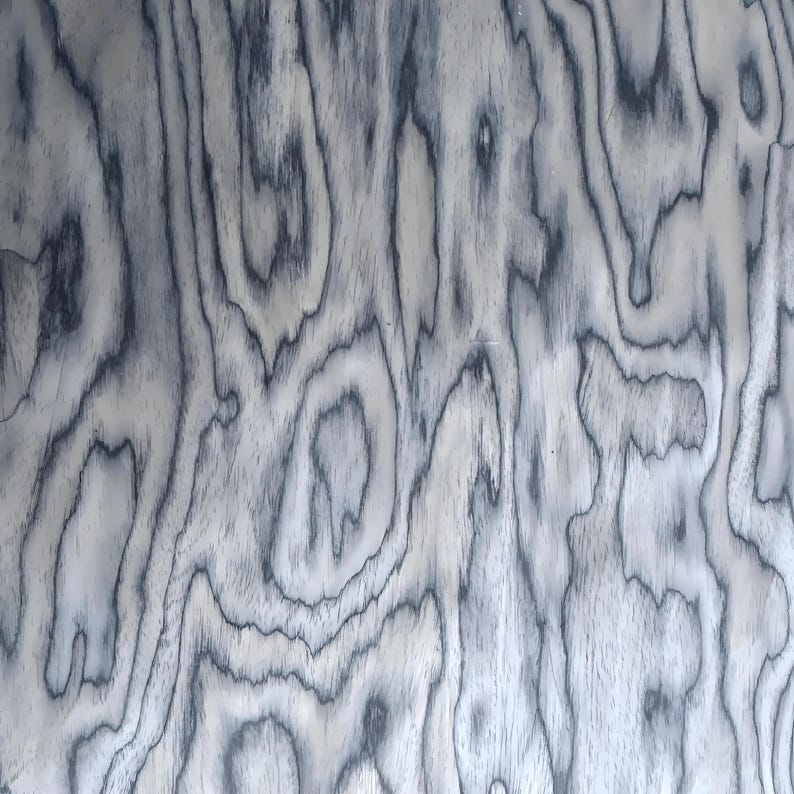 May include: Close-up of a wood grain pattern with a swirling, organic design. The wood features a light grey base with dark grey, almost black, swirling lines that create a textured appearance. The grain pattern is consistent throughout the image.