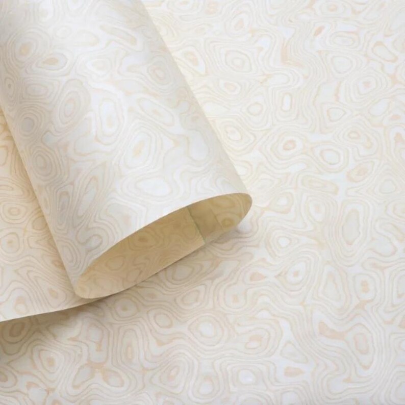 May include: A roll of cream-coloured wallpaper with a unique, organic pattern resembling wood grain. The design features swirling, light brown lines against a white background. The paper is partially unrolled, revealing the pattern and texture.