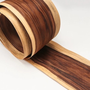 Może przedstawiać: Zbliżenie na rolkę forniru drewnianego. Fornir ma bogate, ciemnobrązowe usłojenie z jaśniejszymi brązowymi krawędziami. Drewno jest zakrzywione, odsłaniając warstwy i naturalne wzory drewna.