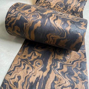 May include: A roll of decorative paper with a swirling pattern in shades of brown and black. The paper has a wood grain appearance, with dark and light areas creating a marbled effect. The paper is partially rolled, revealing the pattern.