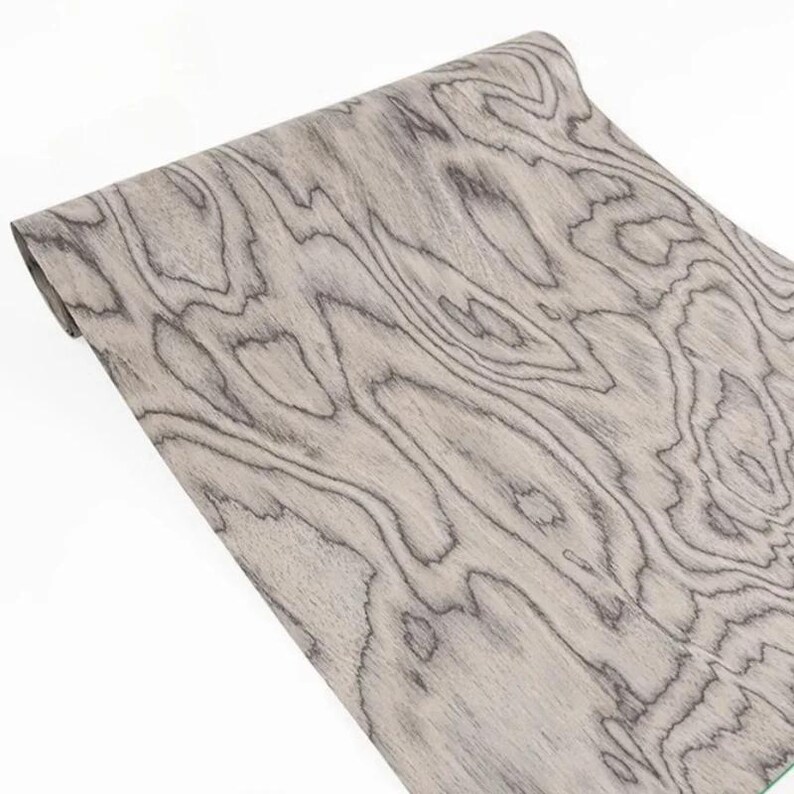 K&ouml;nnte beinhalten: Eine aufgerollte Holzfurnierplatte mit hellgrauer Basis und dunkelgrauen Maserungsmustern. Die Holzmaserung hat ein nat&uuml;rliches, flie&szlig;endes Design. Das Furnier ist wahrscheinlich f&uuml;r M&ouml;bel- oder Dekorationsprojekte bestimmt.