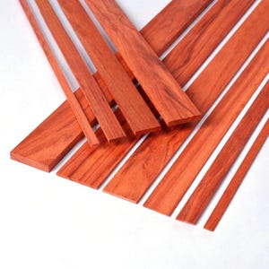 May include: A collection of reddish-brown wooden planks of varying widths and lengths. The planks have a visible wood grain pattern and are arranged on a white surface. The wood appears to be freshly cut and ready for use in woodworking projects.
