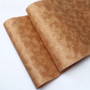 May include: Two sheets of wood veneer with a swirling, organic grain pattern. The veneer has a warm, light brown colour, with darker brown details creating a natural, textured appearance. The sheets are slightly overlapping, showcasing the detailed wood grain.