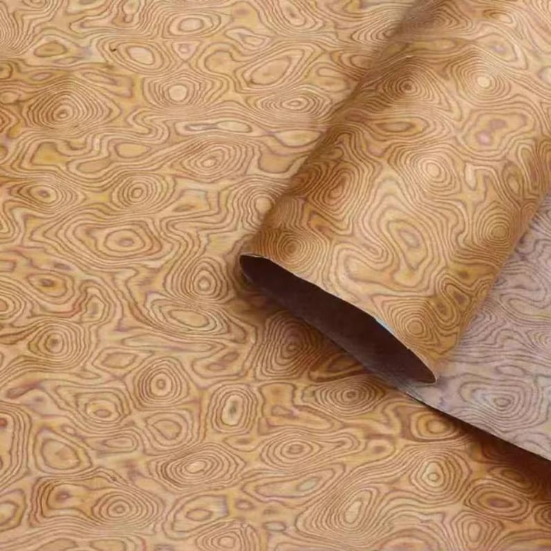 May include: A roll of wood veneer displaying a distinctive, swirling grain pattern in shades of tan and brown. The veneer is partially unrolled, revealing the interior. The pattern resembles topographical maps, creating a natural, organic aesthetic.