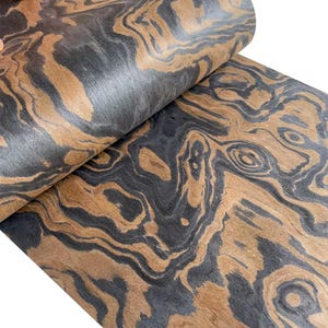 May include: Close-up of a roll of wood veneer with a swirling pattern. The veneer features a combination of dark gray and warm brown tones, creating a natural, organic aesthetic. The wood grain is visible, adding texture and depth to the material.