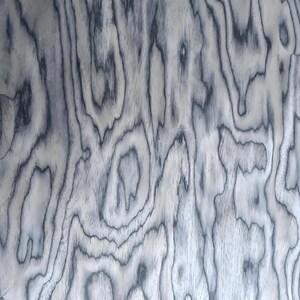 Puede incluir: Primer plano de un patr&oacute;n de vetas de madera con un dise&ntilde;o arremolinado y org&aacute;nico. La madera presenta una base gris claro con l&iacute;neas grises oscuras, casi negras, que crean una apariencia texturizada. El patr&oacute;n de vetas es consistente.