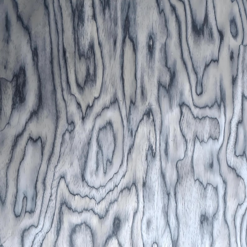 Puede incluir: Primer plano de un patr&oacute;n de vetas de madera con un dise&ntilde;o arremolinado y org&aacute;nico. La madera presenta una base gris claro con l&iacute;neas grises oscuras, casi negras, que crean una apariencia texturizada. El patr&oacute;n de vetas es consistente.