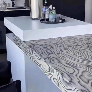 K&ouml;nnte beinhalten: Moderne K&uuml;cheninsel mit wei&szlig;er Arbeitsplatte und einer grau-wei&szlig;en Holzmaserung. Ein schwarzes Tablett enth&auml;lt Wein-, Wasserflaschen und Gl&auml;ser. Eine gro&szlig;e Vase steht auf der Arbeitsplatte.