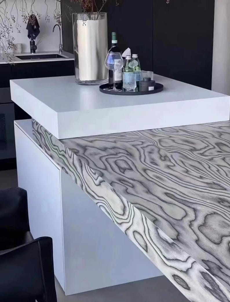 K&ouml;nnte beinhalten: Moderne K&uuml;cheninsel mit wei&szlig;er Arbeitsplatte und einer grau-wei&szlig;en Holzmaserung. Ein schwarzes Tablett enth&auml;lt Wein-, Wasserflaschen und Gl&auml;ser. Eine gro&szlig;e Vase steht auf der Arbeitsplatte.