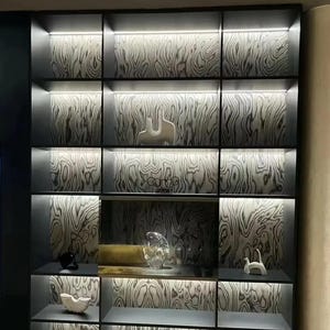 Puede incluir: Un moderno mueble expositor empotrado con m&uacute;ltiples estantes e iluminaci&oacute;n integrada. Los paneles traseros presentan un patr&oacute;n de vetas de madera. Varios objetos decorativos, incluyendo esculturas y piezas de vidrio, se exhiben en los estantes.
