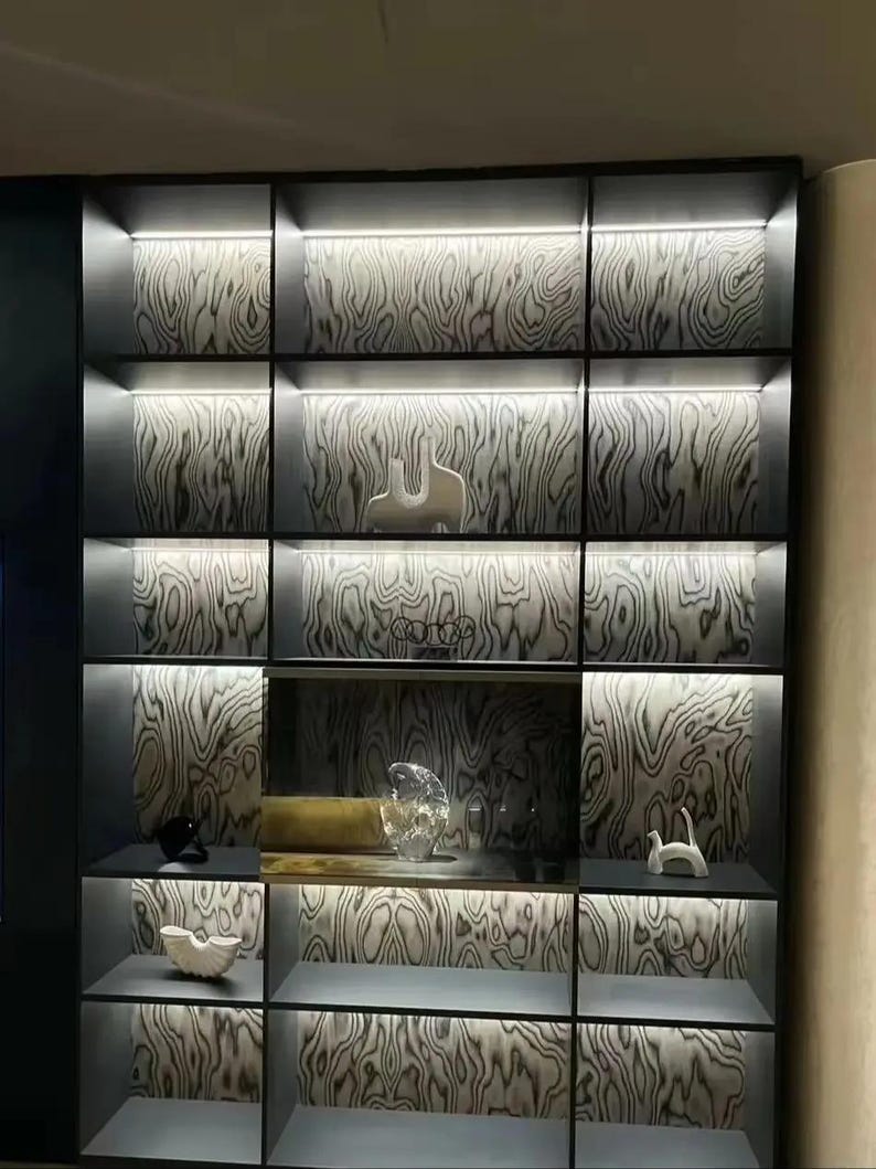 Puede incluir: Un moderno mueble expositor empotrado con m&uacute;ltiples estantes e iluminaci&oacute;n integrada. Los paneles traseros presentan un patr&oacute;n de vetas de madera. Varios objetos decorativos, incluyendo esculturas y piezas de vidrio, se exhiben en los estantes.