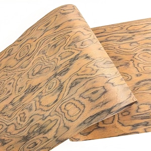 Könnte beinhalten: Zwei Furnierholzplatten mit einem einzigartigen, wirbelnden Maserungsmuster. Das Holz hat eine hellbraune Grundfarbe mit dunkelgrauen und schwarzen Äderungen, wodurch ein natürliches, organisches Design entsteht. Die Platten sind teilweise gerollt und zeigen die Flexibilität des Materials.