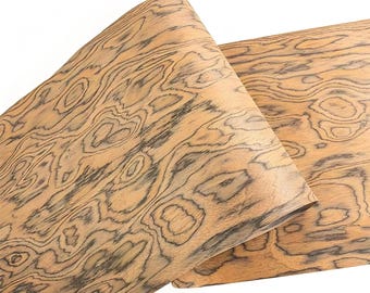 May include: Two sheets of wood veneer with a unique, swirling grain pattern. The wood has a light brown base color with dark gray and black veining, creating a natural, organic design. The sheets are partially rolled, showcasing the flexibility of the material.