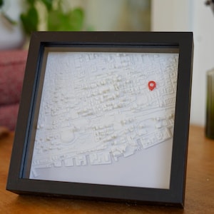 May include: A square black frame containing a white three-dimensional relief map of a city street grid. The map features raised building blocks and a single small red location pin marker placed on a specific intersection within the urban layout.