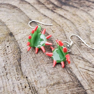 Pode incluir: Um par de brincos de vidro verde e vermelho com um design de espinhos. Os brincos estão sobre uma superfície de madeira.