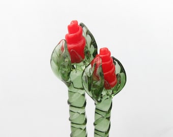 May include: Two glass rose sculptures. Each sculpture features a red rose bud surrounded by green leaves. The stems are green with a spiral design. The background is white.
