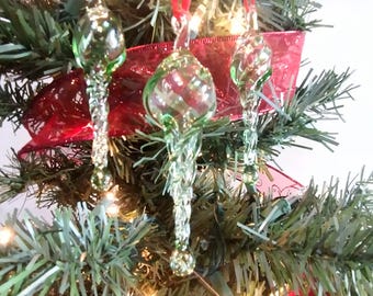 May include: Three green glass Christmas baubles shaped like icicles hanging on a Christmas tree. The baubles are clear glass with a green tint and have a swirl design.