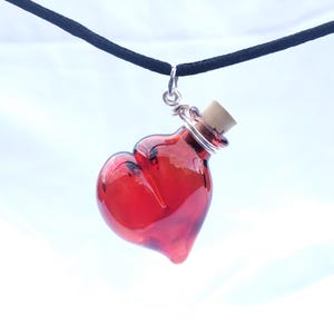 May include: A red glass heart-shaped bottle with a cork stopper hangs from a black cord necklace. The bottle is filled with a clear liquid.