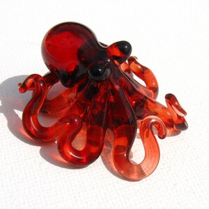 May include: A handcrafted, red glass octopus figurine with black eyes. The octopus has eight curled tentacles and a rounded head. The glass is translucent, allowing light to pass through, and is set against a white background.